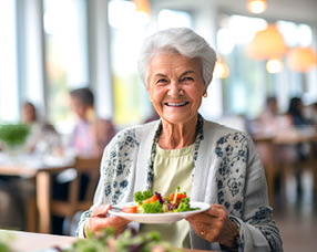 An elderly woman is sitting at a table with a plate of food in front of her. She is smiling and appears to be enjoying her meal. There are other people in the background, and a potted plant is visible in the room. AI generated content