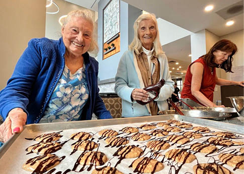 Two women are standing in a kitchen, smiling and preparing food. They are surrounded by a variety of desserts, including cookies and cakes. The women are using a tray to hold the desserts, and there are several bowls and cups nearby. The kitchen is well-equipped with an oven and a sink, and a spoon can be seen resting on a surface. AI generated content