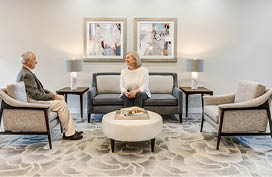 Two people are sitting in a living room, with a couch and chairs arranged around a coffee table. The room is decorated with pictures on the walls and a rug on the floor. AI generated content
