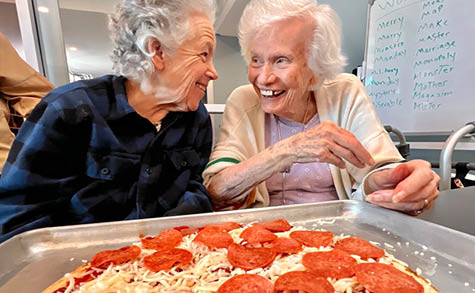 Two older women are sitting at a table with a large pepperoni pizza in front of them. AI generated content